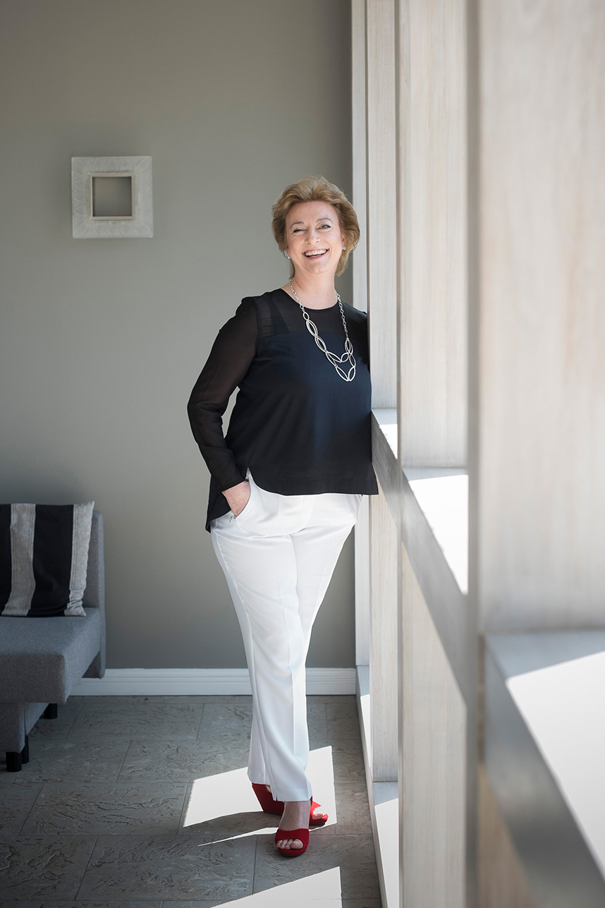 Marion Bevington in a sheer black top, white trousers and red wedge shoes, smiling beside a window.