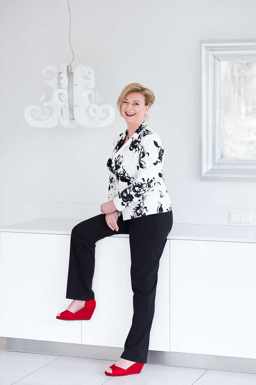 Marion Bevington laughing, leaning against a white sideboard in a black-and-white floral jacket and red wedge shoes.