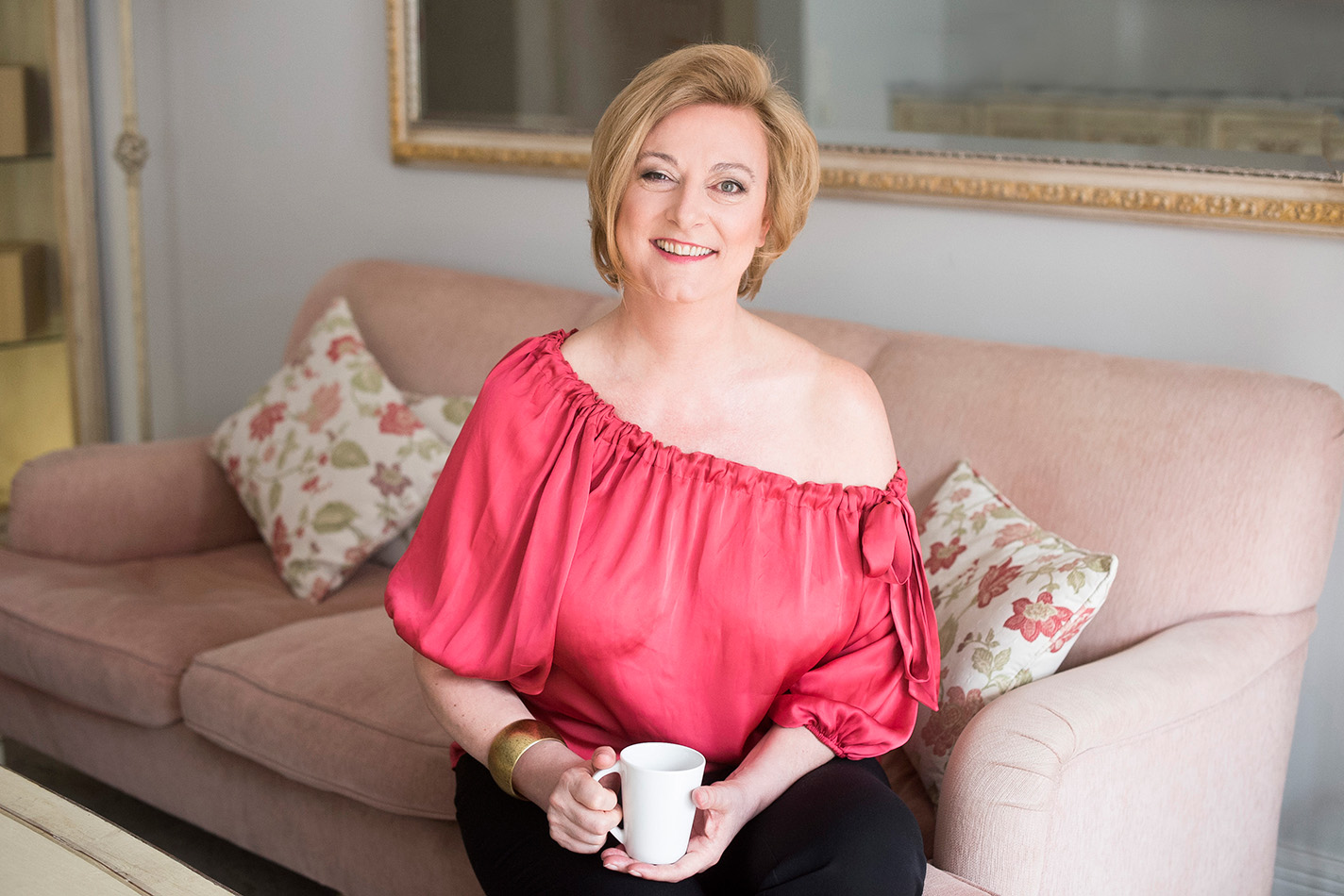 Marion Bevington seated on a pink sofa in a coral off-shoulder top, holding a white mug and smiling warmly.