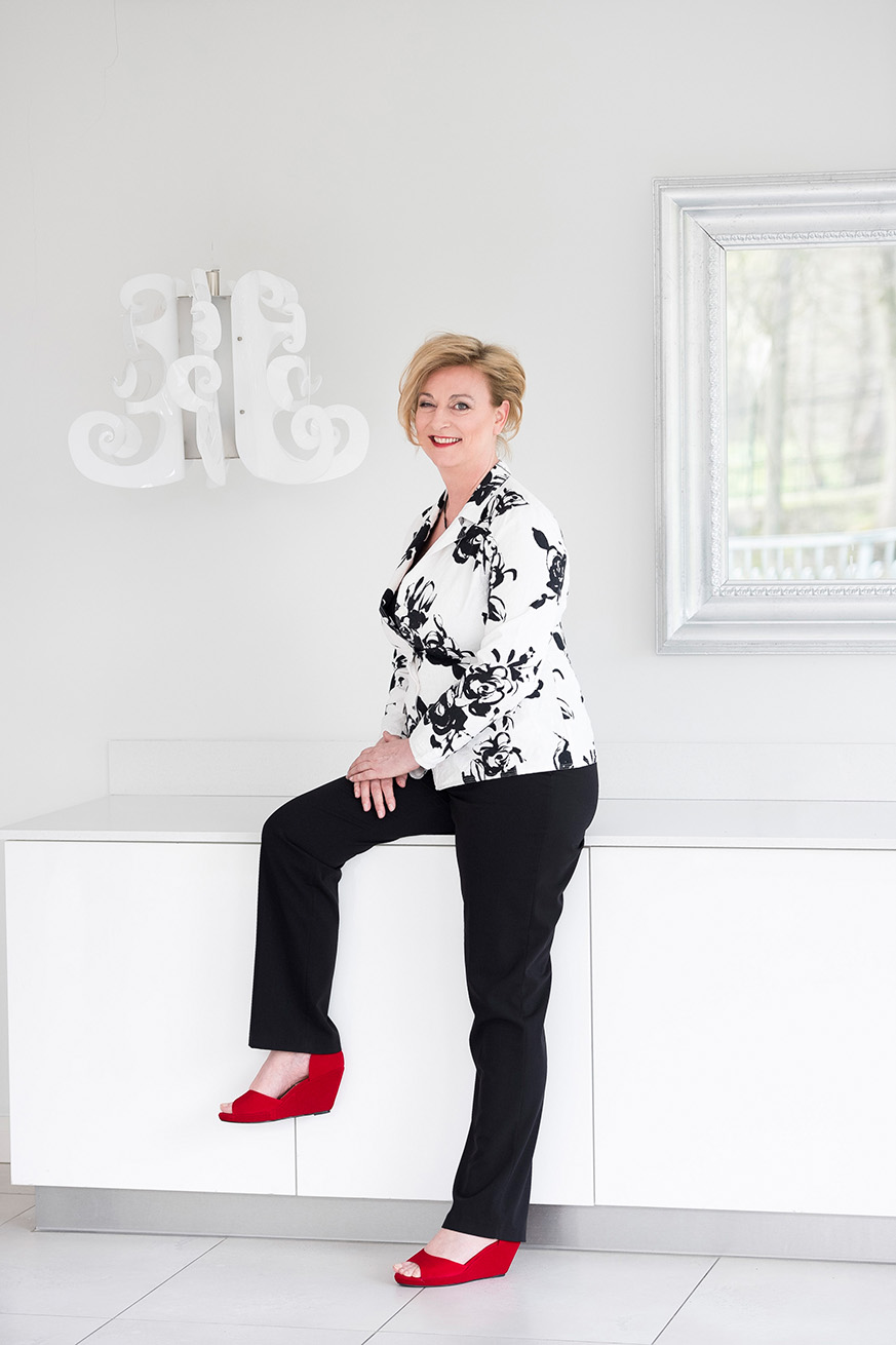 Marion Bevington seated on a white sideboard in a black-and-white floral jacket, smiling at the camera.